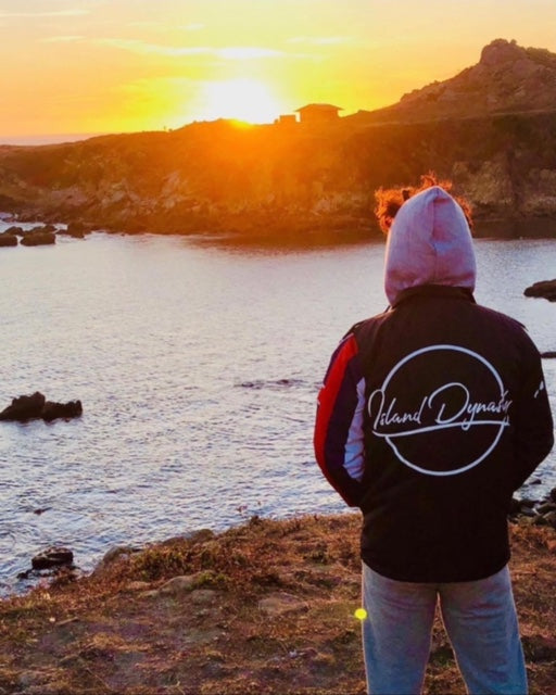 Men's model showing the Back of Island Dynasty Hawaiian Flag Windbreaker displaying the embroidered Island Dynasty Logo on the back standing in front of a sunset. 